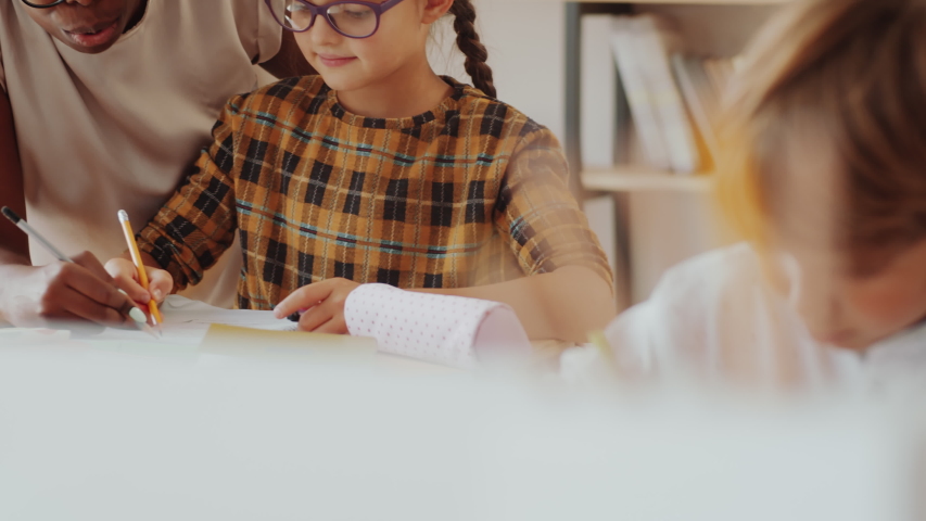 Tilt up shot of happy African American female teacher helping pretty caucasian girl with exercise in workbook, then they looking together at camera and smiling during lesson in primary school Royalty-Free Stock Footage #1057696921