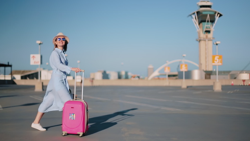 Cinematic shot of happy woman excited about her travel by USA. Slow motion attractive stylish traveler female with pink bag and airport tower on motion background. Fashionable woman walking at sunset Royalty-Free Stock Footage #1057700071