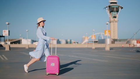 Cinematic shot of happy woman excited about her travel by USA. Slow motion attractive stylish traveler female with pink bag and airport tower on motion background. Fashionable woman walking at sunset
