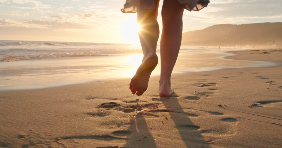Slow motion woman feet walking barefoot by beach at golden sunset leaving footprints in sand. Female tourist on summer vacation in Malibu, California, USA. Woman in beautiful waving dress at sunset | Shutterstock HD Video #1057700104