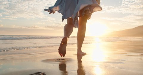 Slow motion woman feet walking barefoot by beach at golden sunset leaving footprints in sand. Female tourist on summer vacation in Malibu, California, USA. Woman in beautiful waving dress at sunset