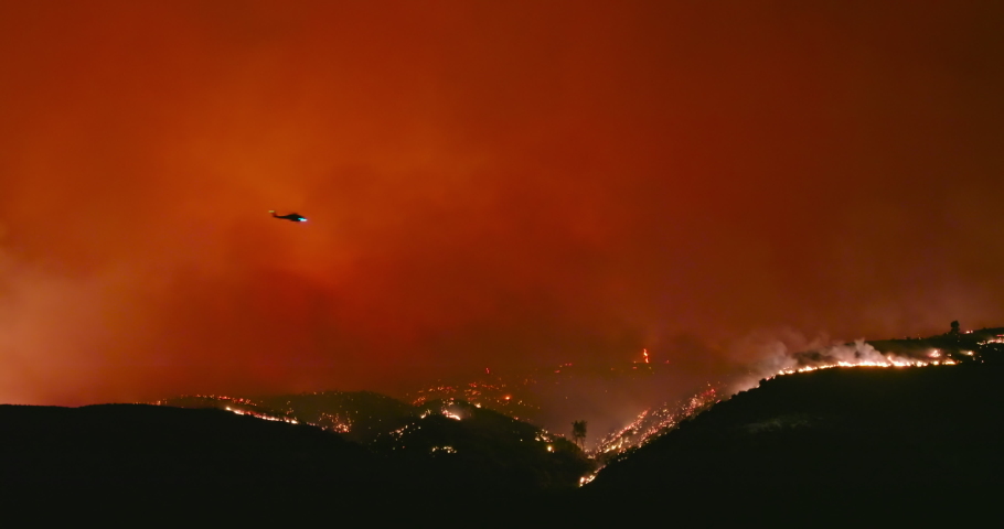 4K epic shot of fire helicopter flying above wildfire. Thick plumes of hot orange smog and red flames rise from forest fire on mountain in Los Angeles suburban. Nature is burning due to climate change