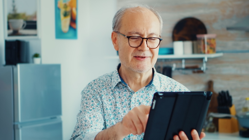 Elderly man using tablet pc in kitchen during breakfast wearing glasses. Elderly person with tablet portable pad PC in retirement age using mobile apps, modern internet online information technology Royalty-Free Stock Footage #1057707586