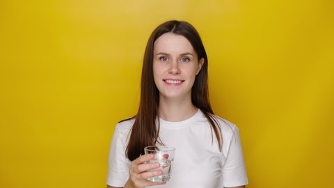 Thirsty young woman holding glass drinks stillwater preventing dehydration, helps maintain normal bowel function and balance of body, isolated on yellow studio background. Healthy lifestyle concept