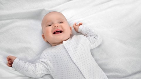 Top view portrait of adorable little baby in sliders lying on white sheet smiling having positive emotion. Happy face of cute toddler enjoying childhood having fun on bed. Medium shot on RED camera