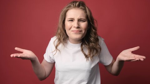 What wrong. Portrait of confused beautiful girl with curly hair, spreads hands sideways, smirks face, feels doubt while makes choice, dressed in casual t shirt. Isolated on red background.