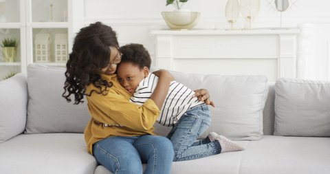 African American young pretty woman in glasses sitting on sofa at home and hugging her cute small daughter. Little girl embracing mother on couch in living room. Motherhood love. Happy safe childhood.