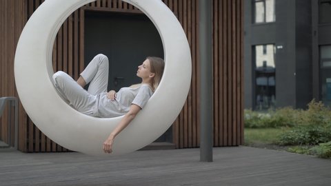 Young woman in white futuristic swing in the yard, gimbal shot. Gimbal shot of young woman having a rest in swings, on background of residential building