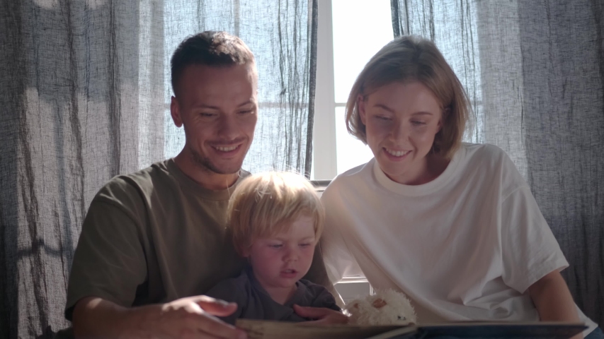 Mom and Dad spend time with 2 year old toddler, show illustrations from the book, sitting together on the bed in room with curtains. Daylight streams in through the curtains. Laying the baby to sleep | Shutterstock HD Video #1057732945