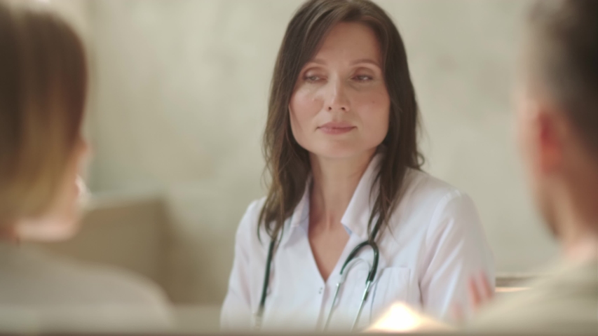 A young woman doctor talks with a couple of patients or relatives (parents) of patients, says the diagnosis, prognosis for treatment. Filmed across the backs of two people in front of a doctor Royalty-Free Stock Footage #1057732960