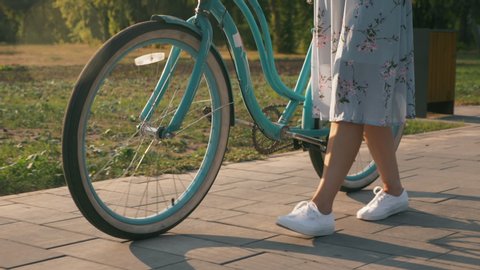 Girl in stylish outfit is walking with vintage retro bike in park in sunshines. Charming girl with bicycle in park at sunset. Bike wheel in sunlight. Cyclist woman is having fun and resting in park