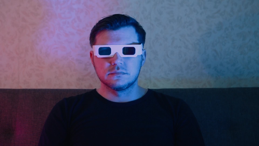 A man sitting on a sofa in stereo glasses emotionally watches TV.