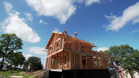 Timelapse view from below of two builders working on the new frame house roof. The blue sky and white clouds are on the background