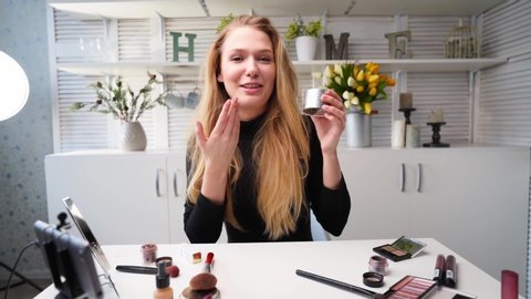 Beauty blogger woman filming daily makeup routine tutorial at camera on tripod. Influencer lady live streaming cosmetics product review in home studio. Vlogger female applies skin powder with brush.