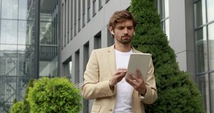 Front view of emotional bearded man using digital tablet outdoors. Handsome businessman in official suit smiling while standing outdoors with modern gadget.