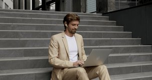 Handsome man in business suit using wireless earphones for communication while typing on laptop. Bearded guy sitting on stairs and working outdoors.