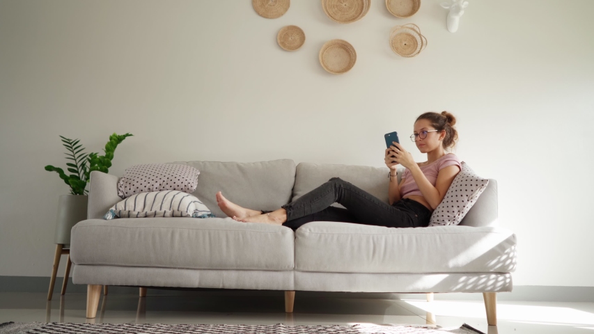 Relaxed young woman using smart phone surfing social media, checking news, playing mobile games or texting messages sitting on sofa. pretty woman spending time at home with smartphone. copy space shot Royalty-Free Stock Footage #1057758982