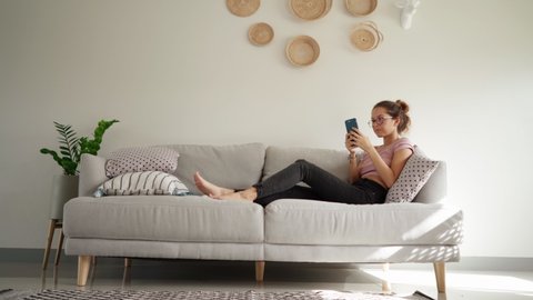 Relaxed young woman using smart phone surfing social media, checking news, playing mobile games or texting messages sitting on sofa. pretty woman spending time at home with smartphone. copy space shot