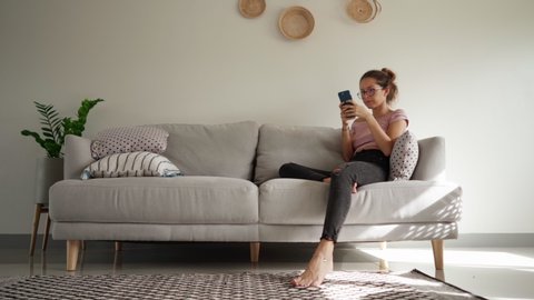 young woman get angry while using smart phone texting messages sitting on sofa, throwing her phone on the sofa