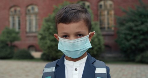 Portrait of kid boy wearing in medical protective mask looking to camera. Child with backpack wearing uniform posing while standing near school. Concept of children, education and virus