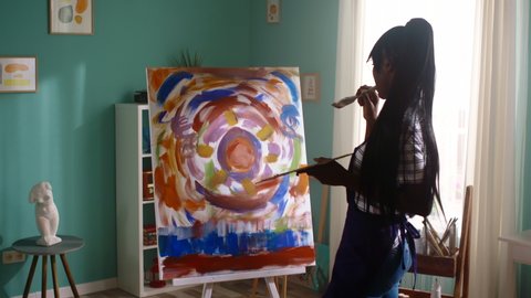 Young black woman is standing in front of beautiful abstract painting, created on canvas with bright saturated acrylic dyes, depiction of symbolic sun that means joy and positive energy, Slow motion.