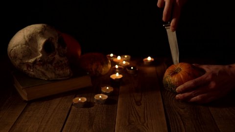 Carving top of a pumpkin against background of a skull and candlelight.. Carving jack-o-lantern on a wooden table in orange light of candles. Theme feast of all saints in autumn on harvest of pumpkins Stock Video