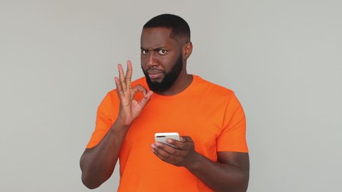 Happy fun african man in orange basic casual t-shirt hold talk using mobile cell phone chatting just found out great big win news doing ok okay hand gesture isolated on gray color background in studio