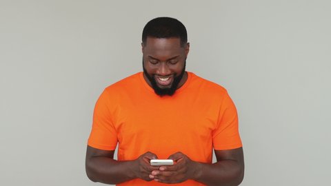 Young shocked happy win bearded african american man guy male 20s in orange basic casual t-shirt look surprised say wow hold using mobile cell phone isolated on grey color gray background in studio