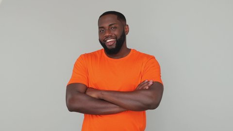 Young bearded african american guy 20s in orange basic casual t-shirt look at camera with charming smile with folded crossed hands on chest blink wink eye isolated on grey color gray background studio