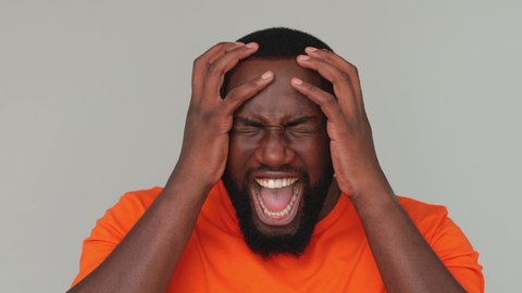 Close up young mad angry displeased african man in orange basic casual t-shirt looking at camera screaming swearing expressive gesticulating with hands isolated on grey color gray background in studio