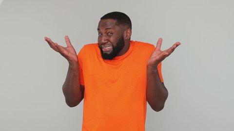 Young fun confused shy shamed bearded african american man guy in orange basic casual t-shirt looking at camera spreading hands say oops i am so sorry isolated on grey color gray background in studio