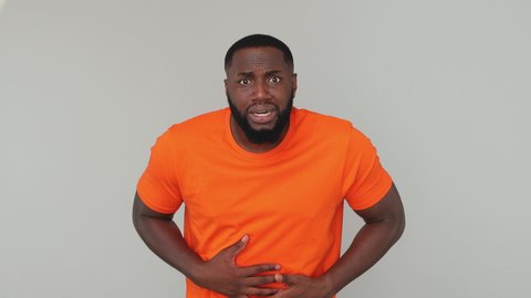 Young sad sick bearded african american man guy male 20s wearing orange basic casual t-shirt look at camera put hands on tummy have stomach ache pain isolated on grey color gray background in studio
