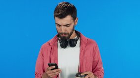 Portrait of young man drinking coffee from paper cup on blue background and looking at phone. He wears headphones. Slow motion video.
