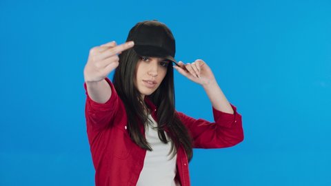 Brunette woman in hat showing middle finger, rude sign of disrespect, making a fuck gesture with resentful resentful look. Indoor studio shot isolated on blue background
