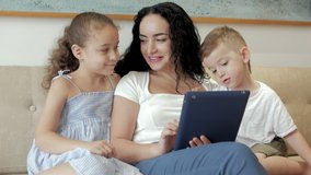 Happy family,mom and cute children watch tablet or laptop,mother plays with children at home, relaxing using tablet, hugging, sitting on the couch,laughing, having fun,enjoying moments of family life.