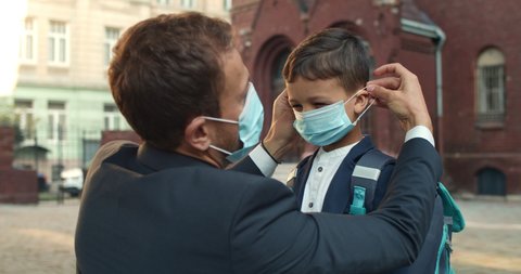 Close up of loving father wearing medical mask on his son face while sending him to school. Boy with backpack giving high five and running while man in suit standing and watching