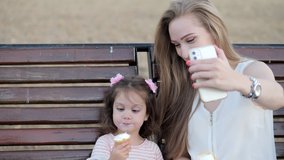 Young mother and her daughter eating ice-cream and talking on a video call on smartphone while sitting on bench in park