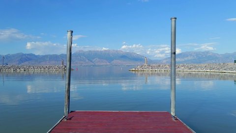 Fixed shot from the end of a red wooden jetty looking out towards the Saratoga Springs Marina entrance with flat water and crystal clear mountains in the background, only a few clouds in the distance.