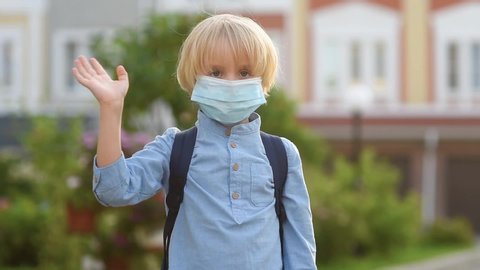 Child wearing face mask going at reopen school after covid-19 quarantine and lockdown. It is new normal for protection and prevention while outbreak of coronavirus or flu. Schoolboy waves goodbye.