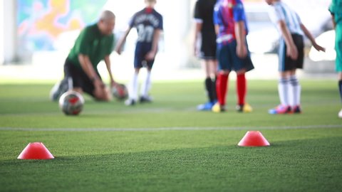 Selective focus to red marker cone is soccer training equipment on green artificial turf with blurry kid players training background. Material for training class of football academy.