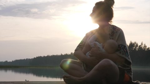 Young mom breastfeeding her baby on the dock of the lake at sunset.Mother baby bonding time.