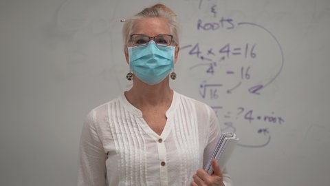 Portrait of smiling mature woman math teacher wearing medical face mask set against a white board with algebra equations in a school classroom.