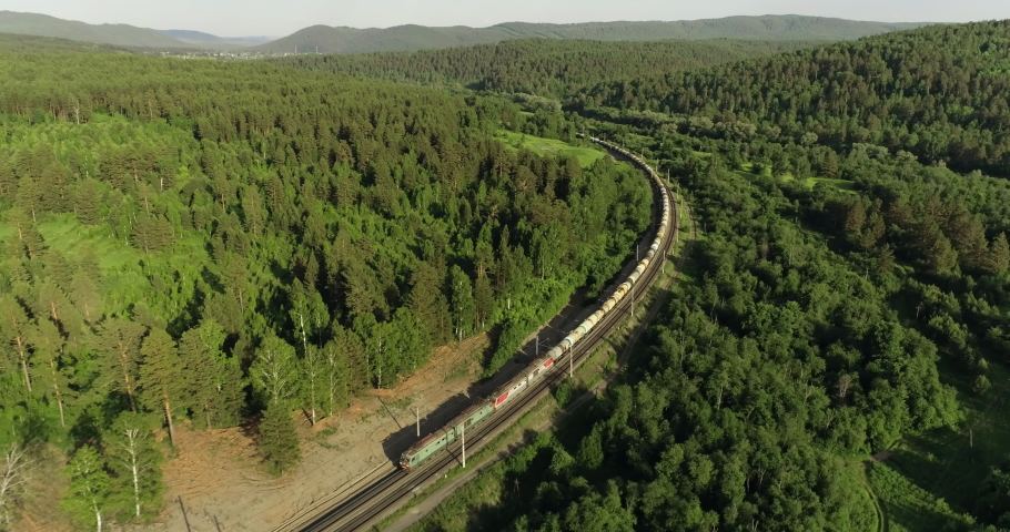 Freight long train carries with oil tank and petrol carriages an electric locomotive by Trans Siberian railways under the rock and near mountain river / Aerial drone wide view at summer sunny day Royalty-Free Stock Footage #1057805971