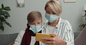 Crop view of ill family using phone whilw sitting on sofa at home. Woman and her teen son in medical masks looking at smartphone screen while watching video. Concept of illness.