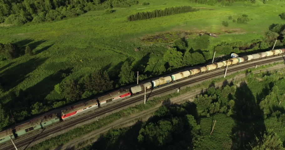 Freight long train carries with oil tank and petrol carriages an electric locomotive by Trans Siberian railways under the rock and near mountain river / Aerial drone wide view at summer sunny day Royalty-Free Stock Footage #1057808758
