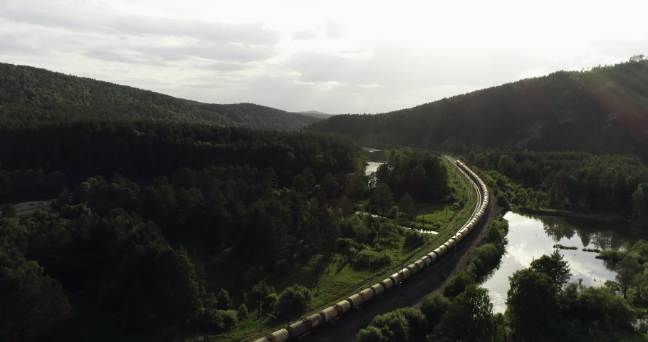 Freight long train carries with oil tank and petrol carriages an electric locomotive by Trans Siberian railways under the rock and near mountain river / Aerial drone wide view at summer sunny day Royalty-Free Stock Footage #1057808761