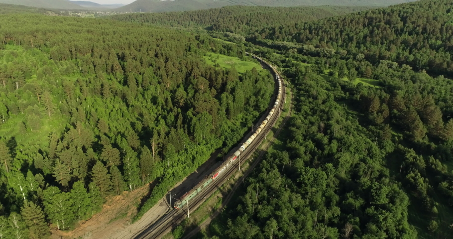 Freight long train carries with oil tank and petrol carriages an electric locomotive by Trans Siberian railways under the rock and near mountain river / Aerial drone wide view at summer sunny day Royalty-Free Stock Footage #1057815715