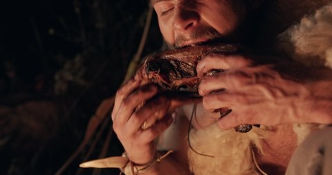 Low angle of hungry male barbarian eating fried meat and looking around in fear while resting near campfire at night. Caveman Neanderthal wildly eats baked meat and looks around to see if there is any
