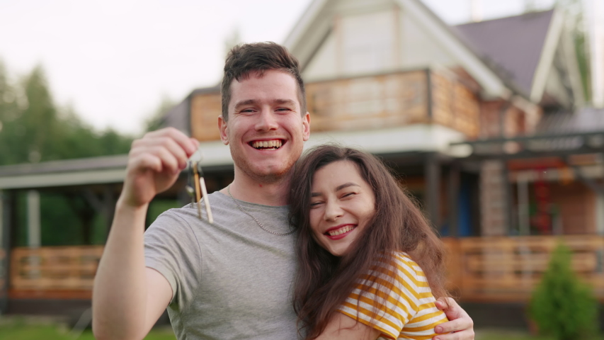 Young married couple or family buy a new house. Happy man and woman or owners shake key in air and hug each other in front of their newly bought home. Excited clients purchasing estate or apartment | Shutterstock HD Video #1057819744