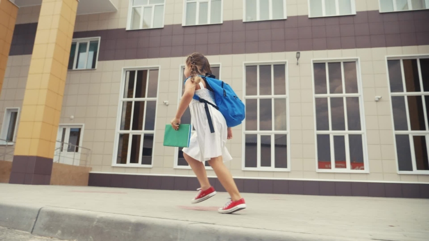 little girl kid with a backpack and a textbook runs hurrying to school. fun education concept. little schoolgirl with a backpack runs to the school building. child running with textbook back view Royalty-Free Stock Footage #1057821346